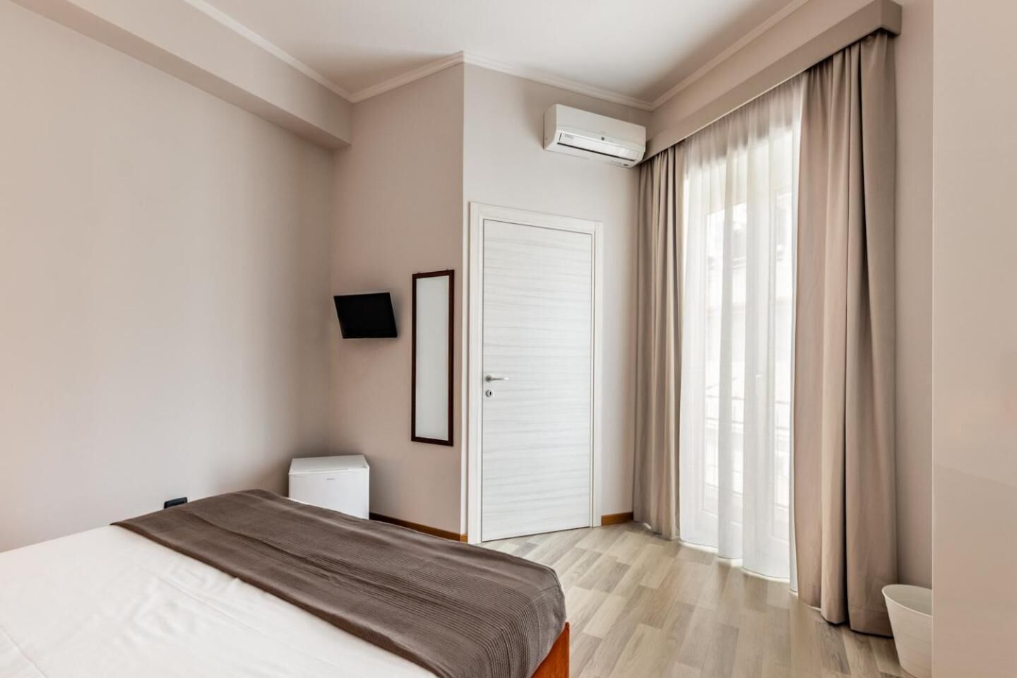 Ett hotellrum med en säng, en tv som är monterad på väggen, en vit garderob och ett fönster med gardiner.