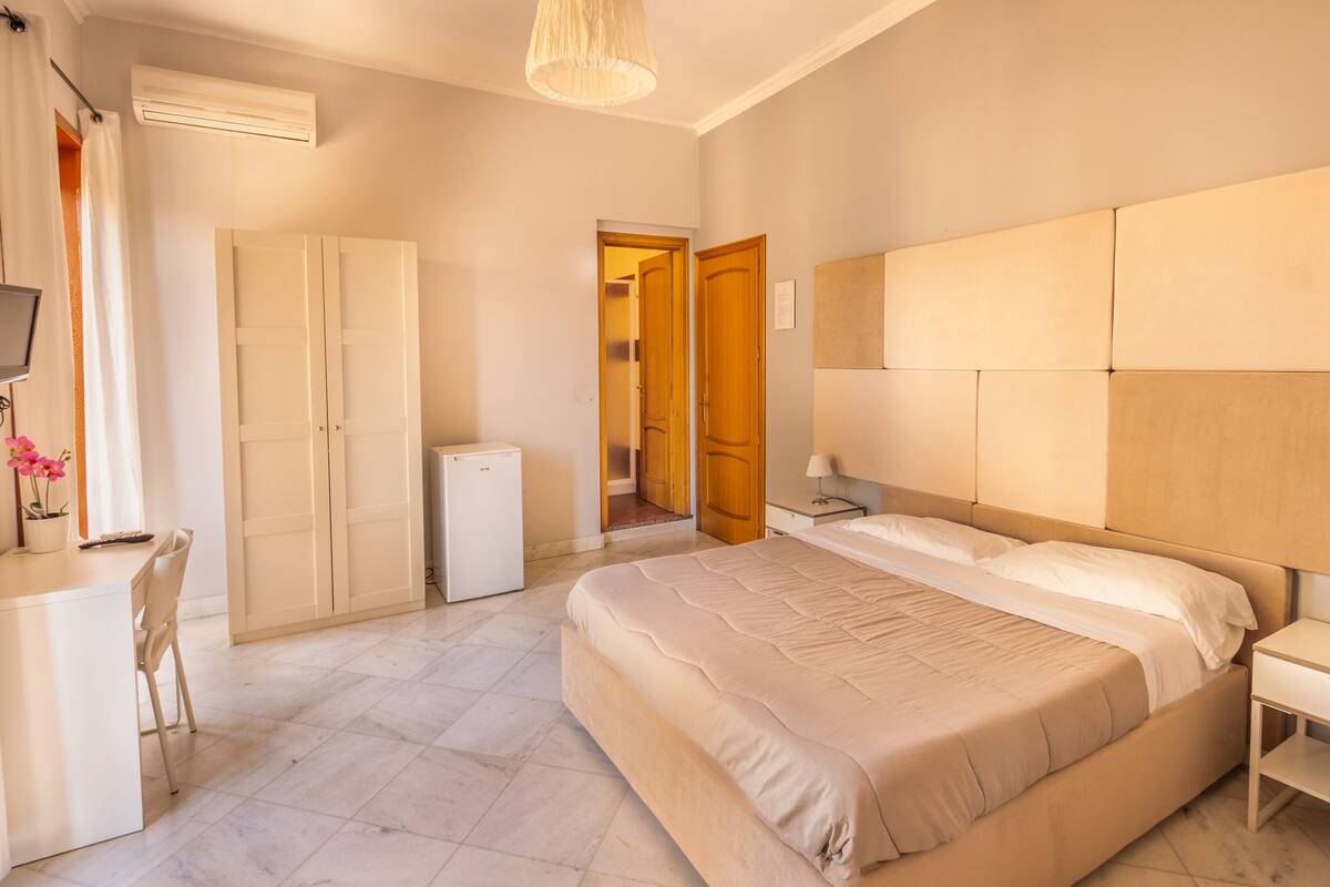 A bedroom with a bed, a desk, a chair, a wardrobe, and a small refrigerator.