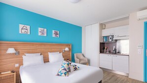 Hotellihuone, jossa on suuri sänky, puinen pääty, sininen korosteseinä, pieni keittiö ja tuoli.