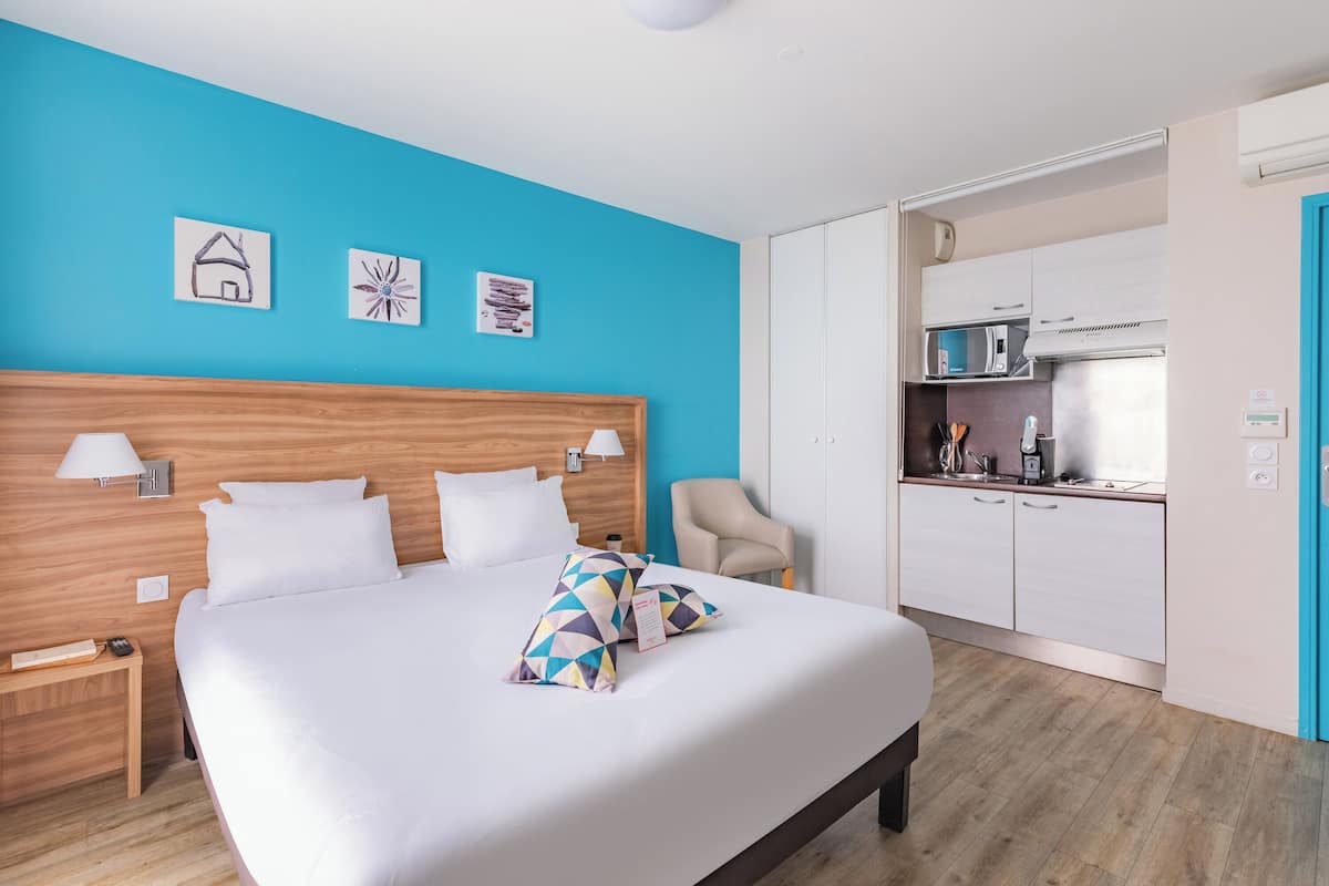 A hotel room with a large bed, a wooden headboard, a blue accent wall, a kitchenette, and a chair.