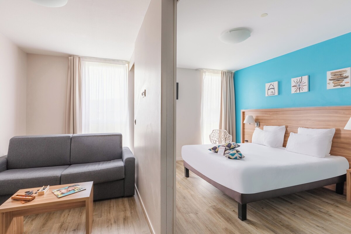 A modern hotel room with a grey sofa, a wooden coffee table, a bed with white bedding, and a blue accent wall.