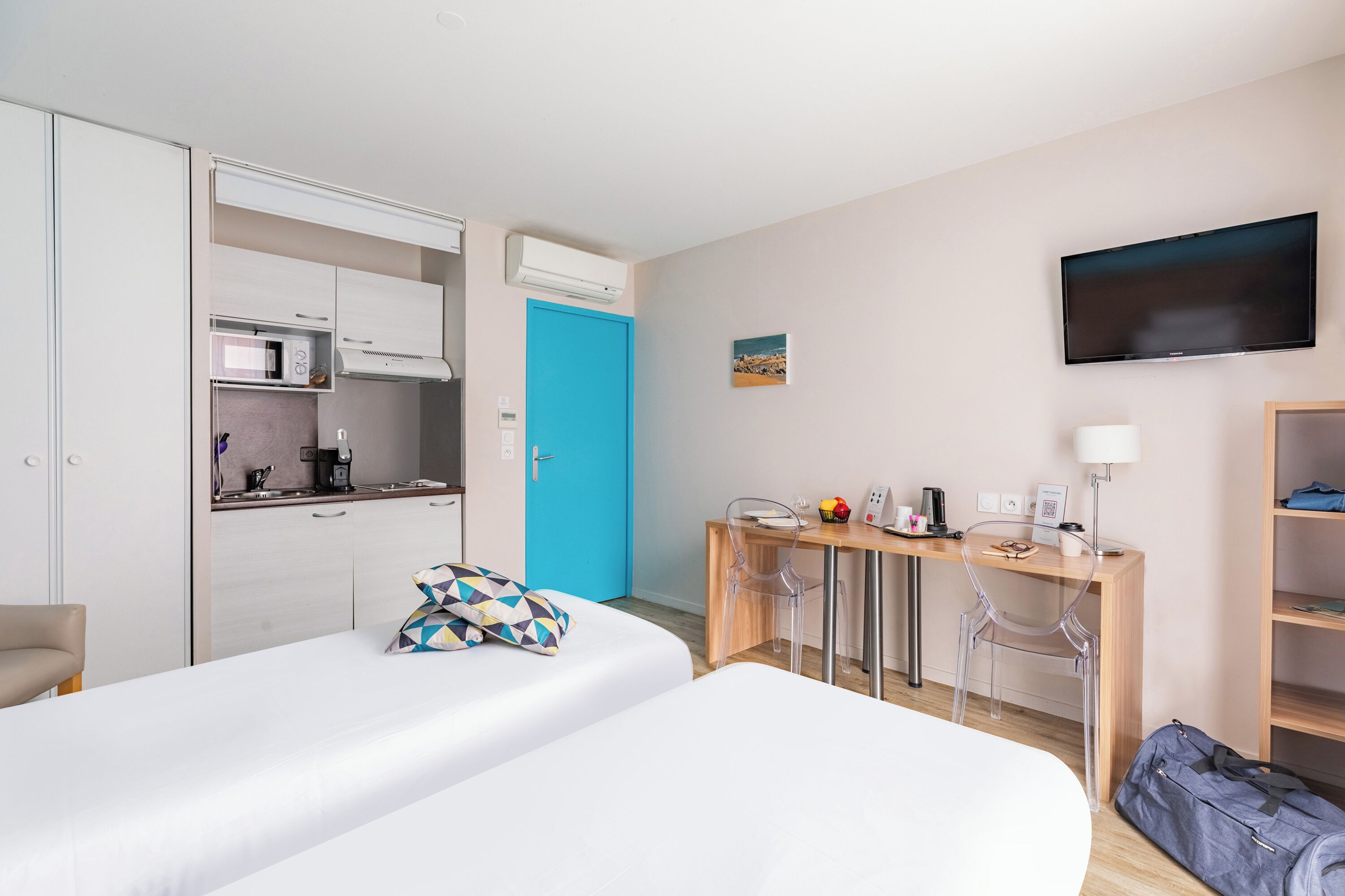 A hotel room with a bed, a desk with a chair, a TV, and a kitchenette.
