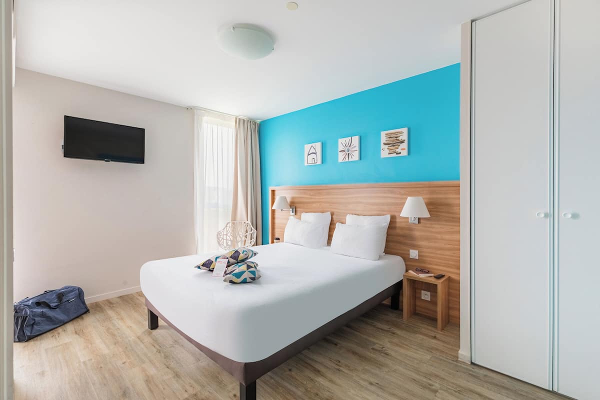 A modern hotel room with a large bed, a flat-screen TV mounted on the wall, and a wooden headboard.