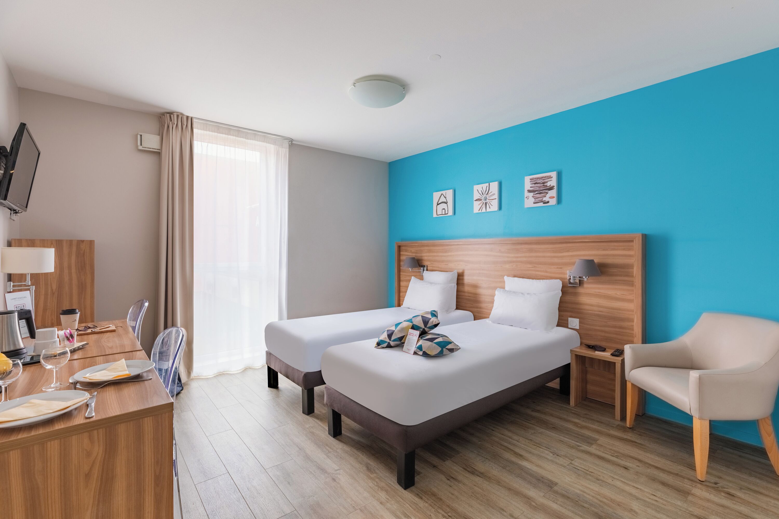 Une chambre d’hôtel avec deux lits, un bureau avec une chaise, une télévision et des œuvres d’art accrochées aux murs.