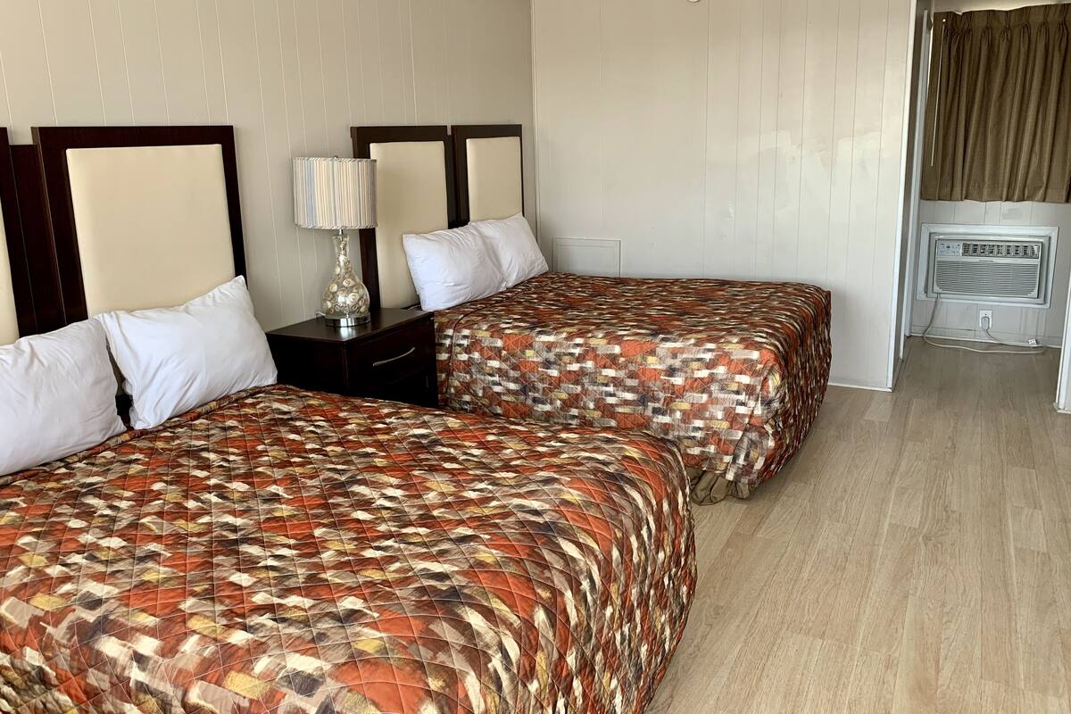 A hotel room with two beds, a nightstand with a lamp, a small table, and a window with curtains.