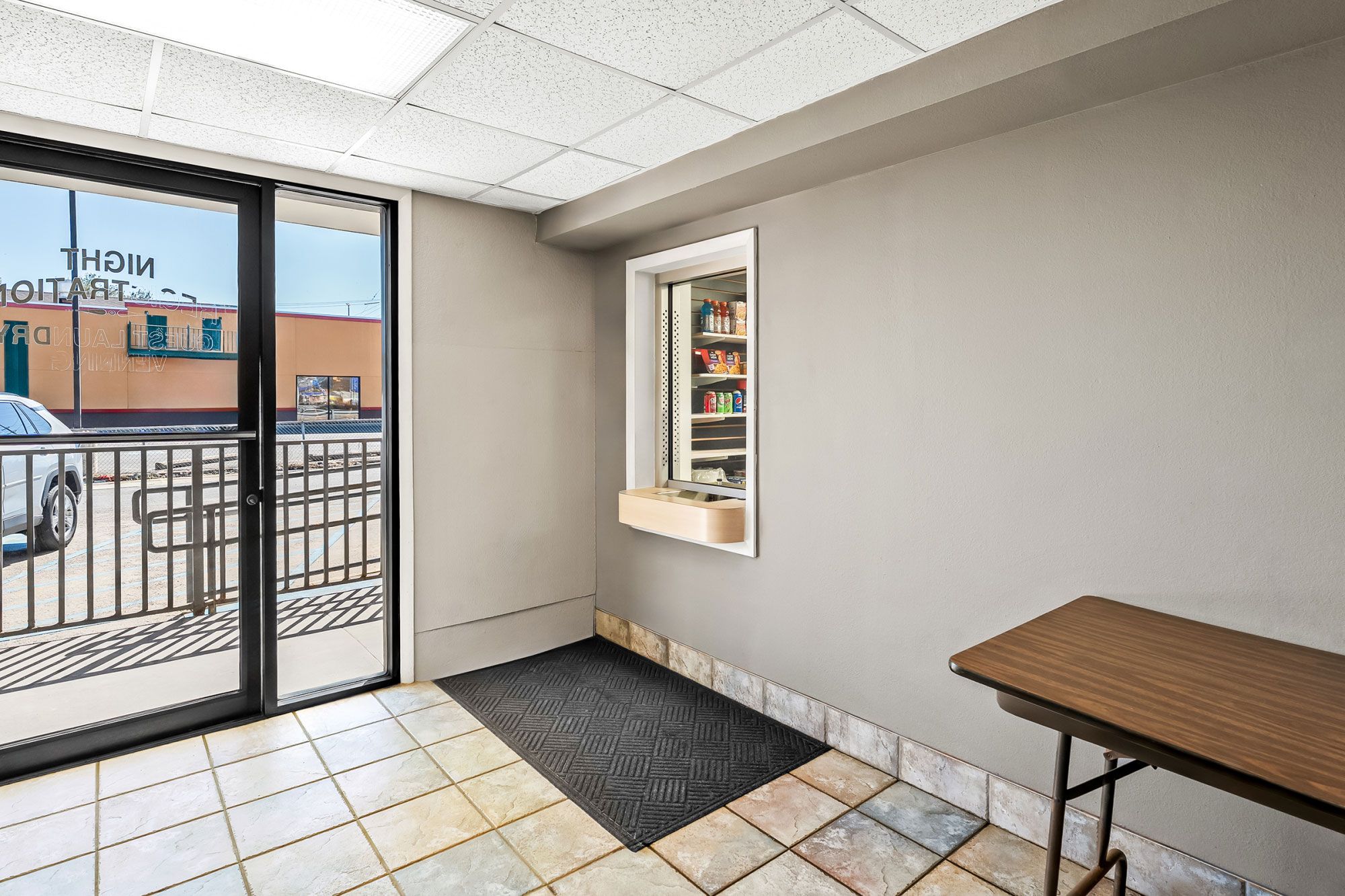 Habitación de hotel con puerta de vidrio corredera, un lavamanos pequeño, un espejo, una mesa de madera y piso de baldosas.