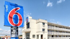 Une enseigne de motel avec un grand chiffre « 6 » et un fond bleu, un bâtiment blanc doté de balcons, et un ciel dégagé.