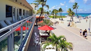 Balcón de hotel con sillones de sol y sombrillas, con vista a una playa con palmeras y gente.