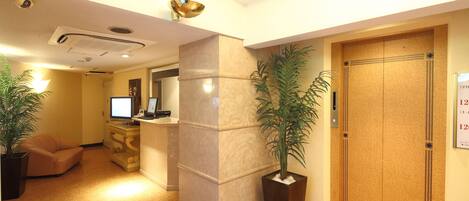 Un vestíbulo de hotel con mostrador de recepción, un pilar grande, una puerta de madera y plantas en macetas.