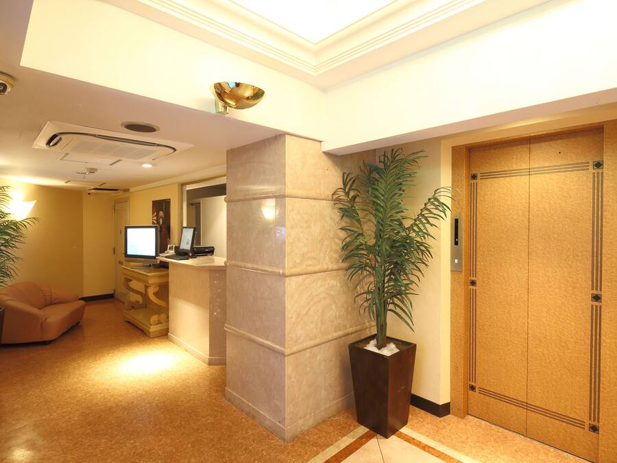 Um saguão de hotel com balcão de recepção, um pilar grande, uma porta de madeira e plantas em vasos.