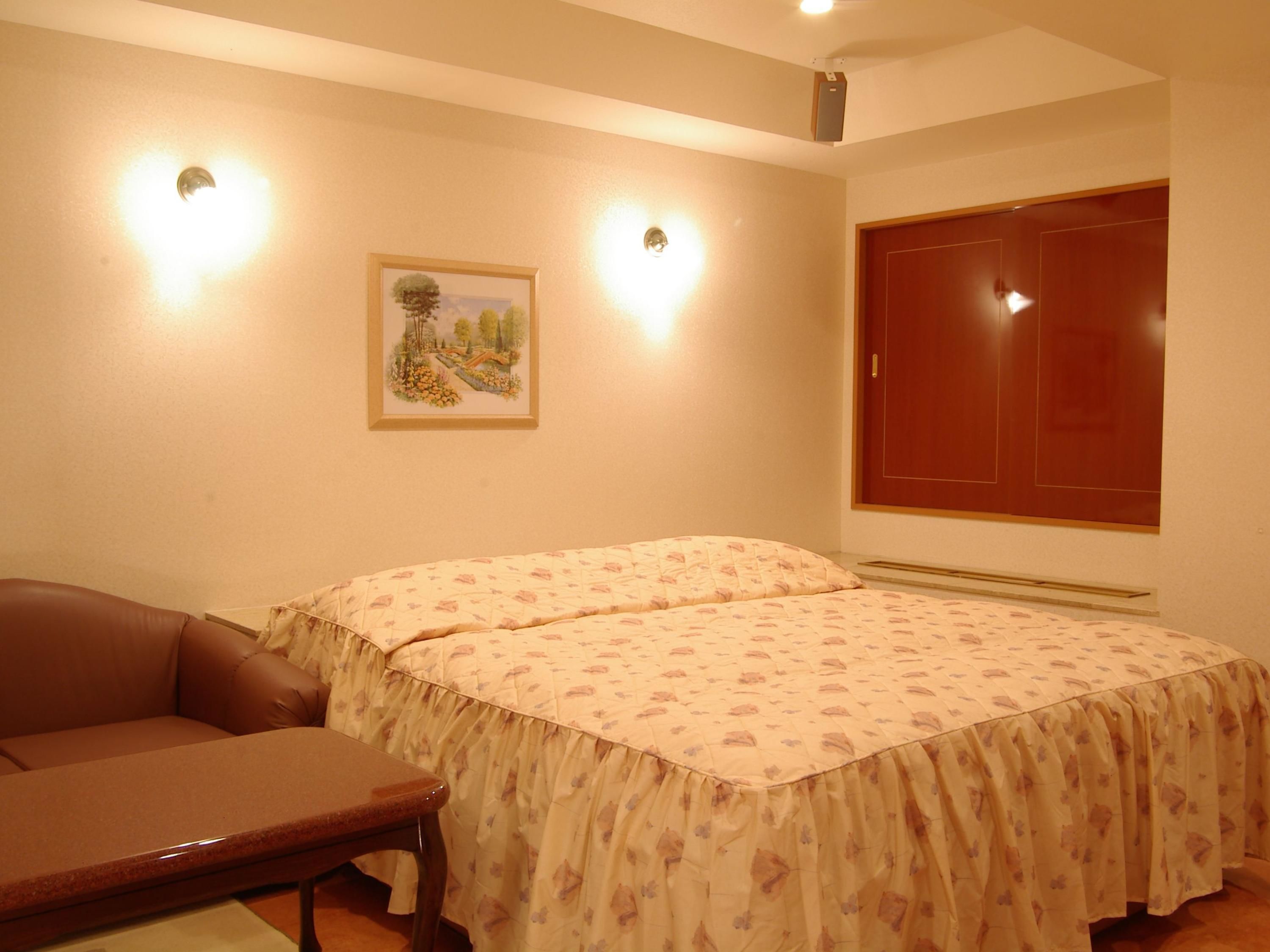 A bedroom with two beds, a wooden wardrobe, a framed picture on the wall, and two wall-mounted lights.