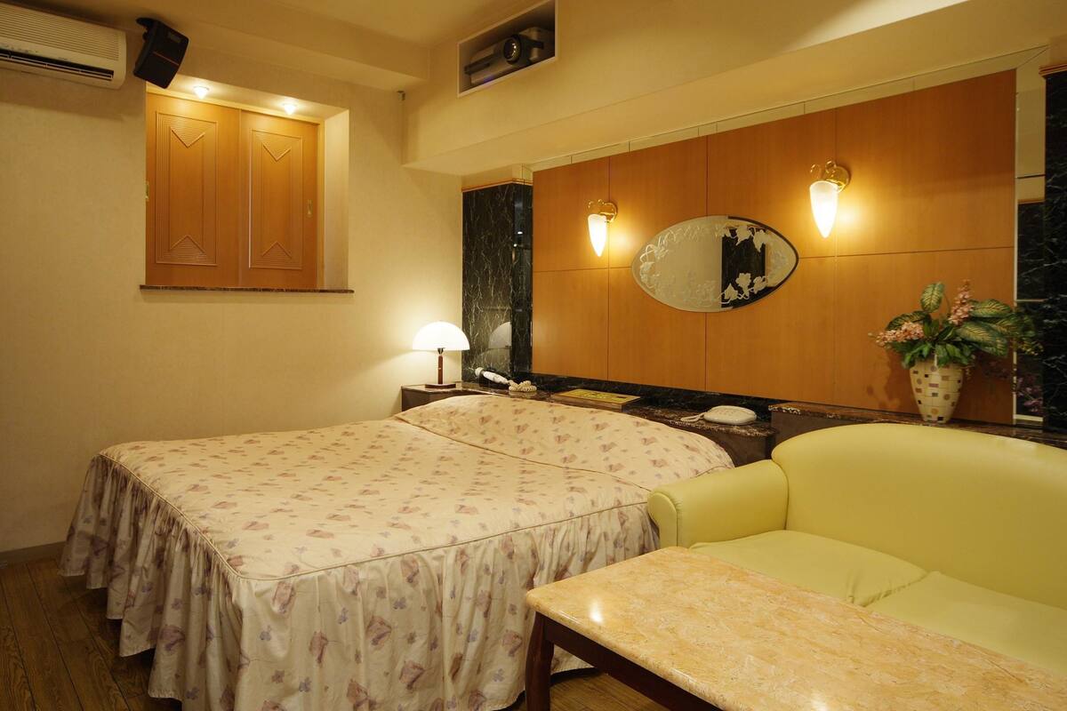 A hotel room with two beds, a sofa, a small table, a mirror, a lamp, a potted plant, and a wall-mounted air conditioner.
