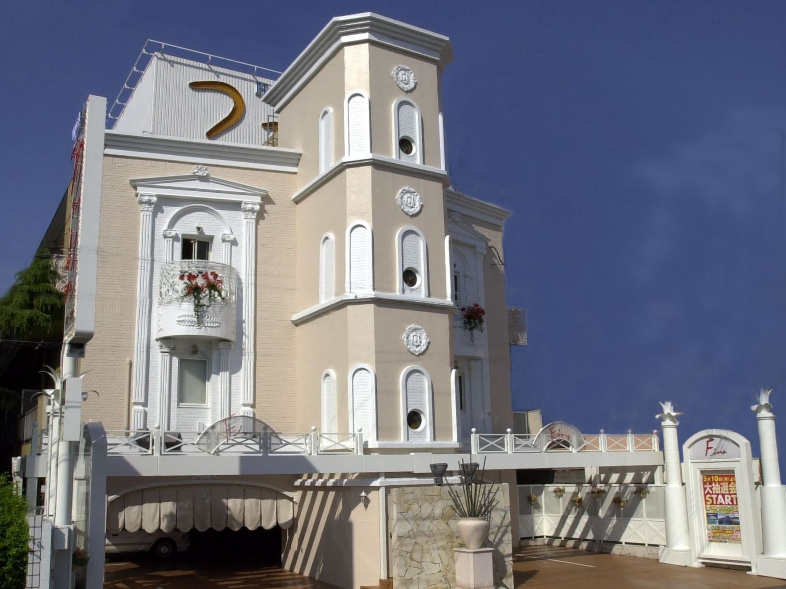 A multi-story building with a white facade, a balcony with flowers, and a large golden 'D' on the roof.