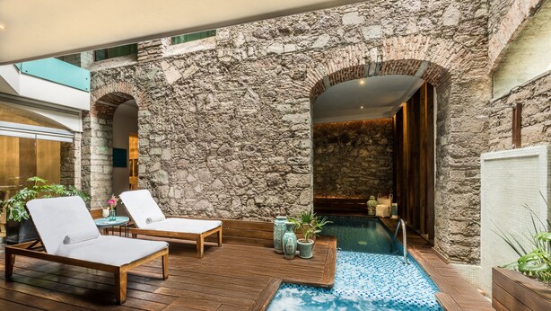 Un espacio al aire libre con paredes de piedra, una terraza de madera, dos sillones de descanso, una piscina pequeña y plantas en macetas.
