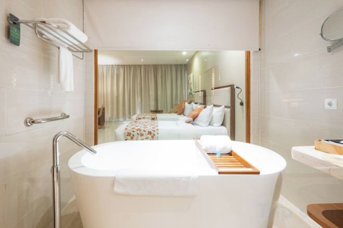 Un baño moderno con una bañera grande, zona de ducha y una habitación visible con cama y cortinas.