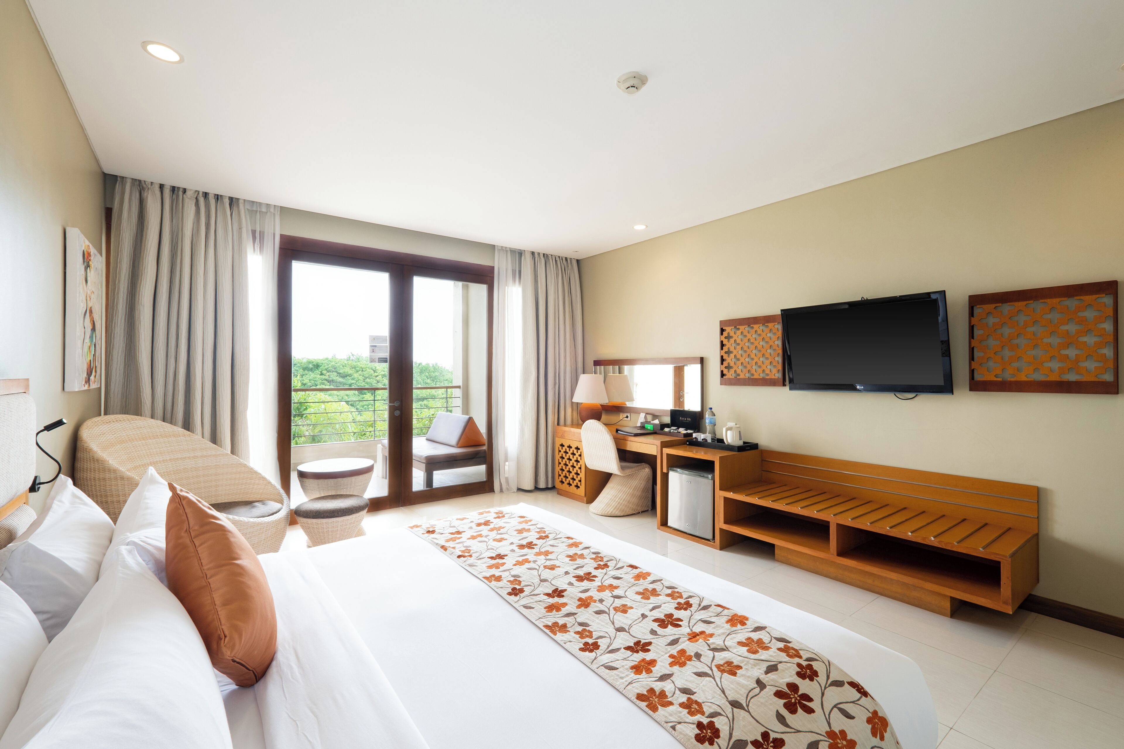 A hotel room with a large bed, a desk with a computer, a television, and a balcony with a view of greenery.
