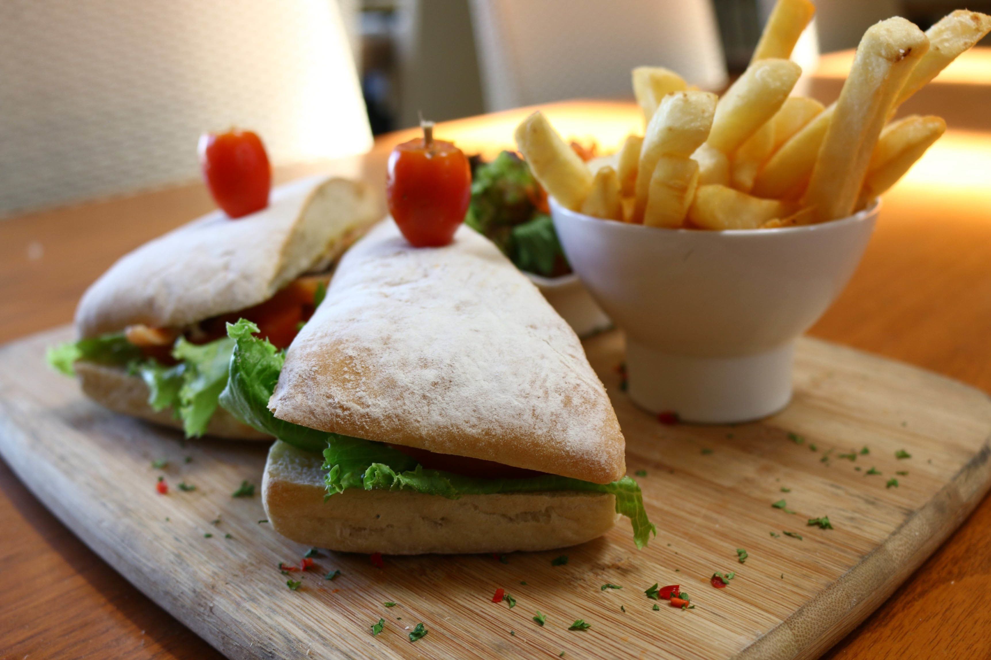 A sandwich with lettuce and a small tomato on top, accompanied by a bowl of fries on a wooden board.