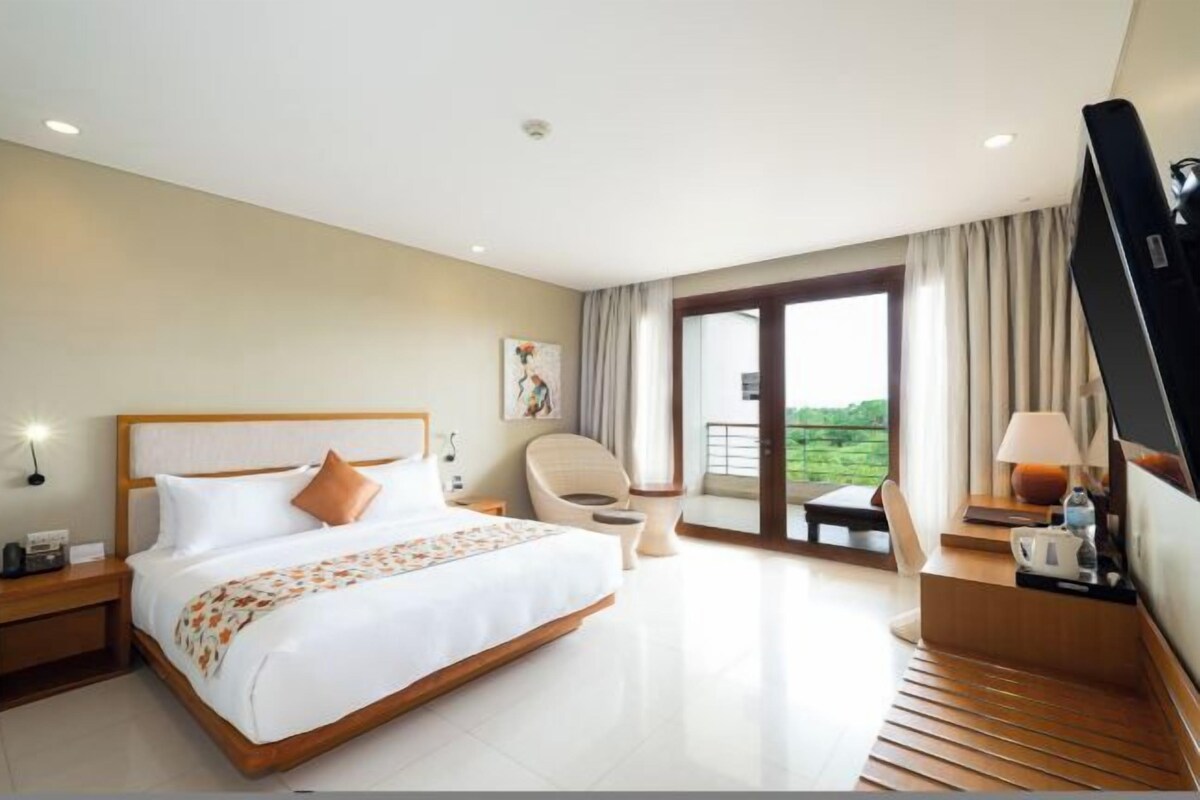 A hotel room with a large bed, a chair, a desk with a TV, and a balcony with a view.
