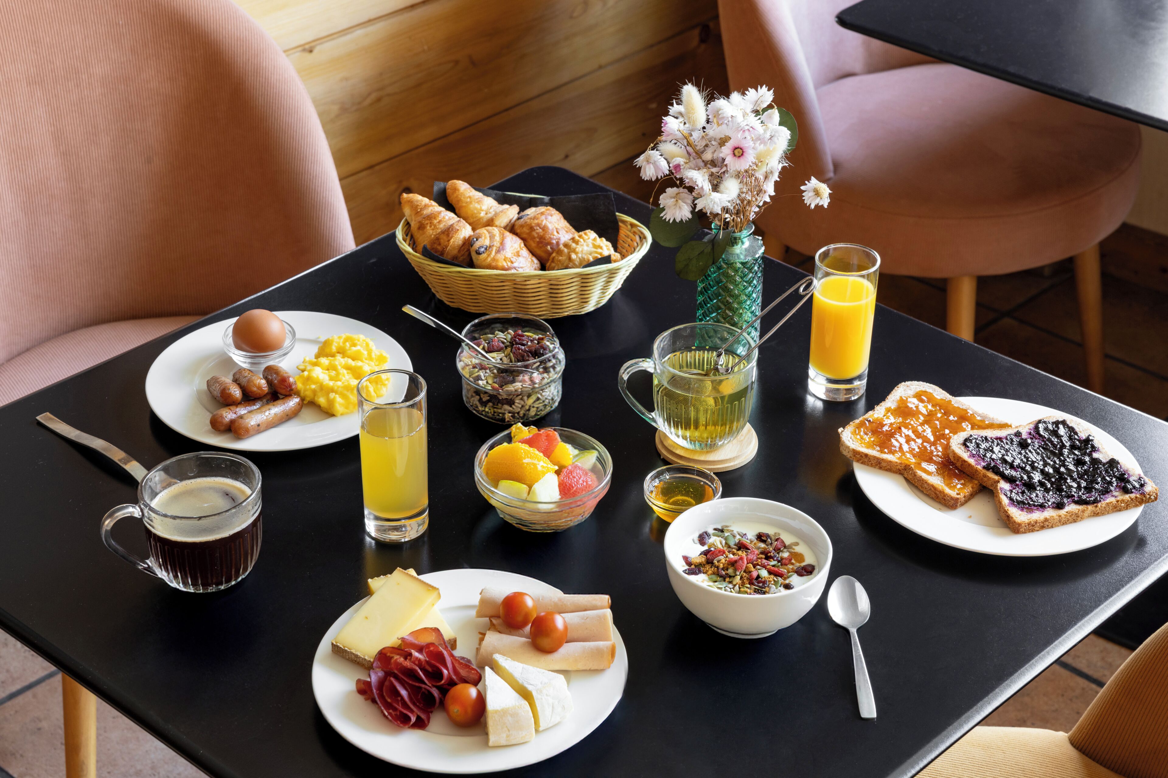 A breakfast table with various food items, a basket of croissants, a vase with flowers, and a glass of orange juice.