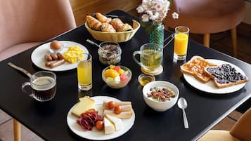 A breakfast table with various food items, a basket of croissants, a vase with flowers, and a glass of orange juice.
