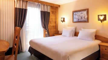 A hotel room with a large bed, wooden headboard, two bedside tables, a nightstand, a wooden chair, and a window with curtains.