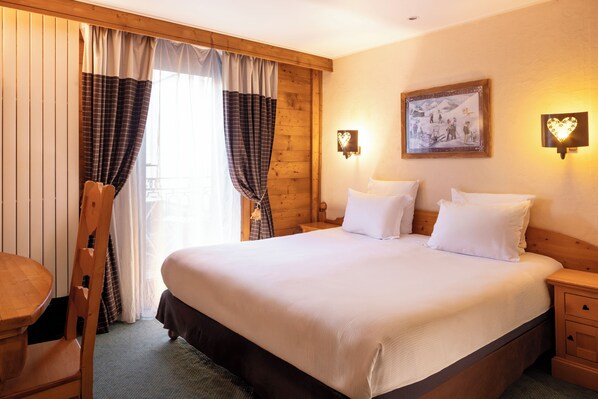 Habitación de hotel con una cama grande, cabecera de madera, dos mesitas de noche, un sillón de madera y una ventana con cortinas.