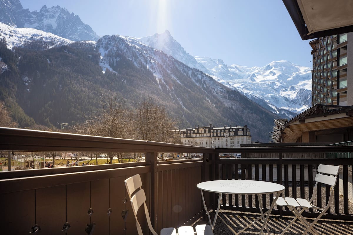 Een balkon met een tafel en stoelen, met uitzicht op een bergachtig landschap met besneeuwde toppen.