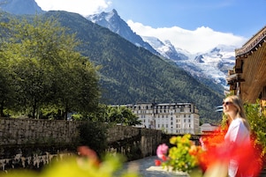 Edificio histórico con fachada de piedra, un río al frente y montañas nevadas al fondo.