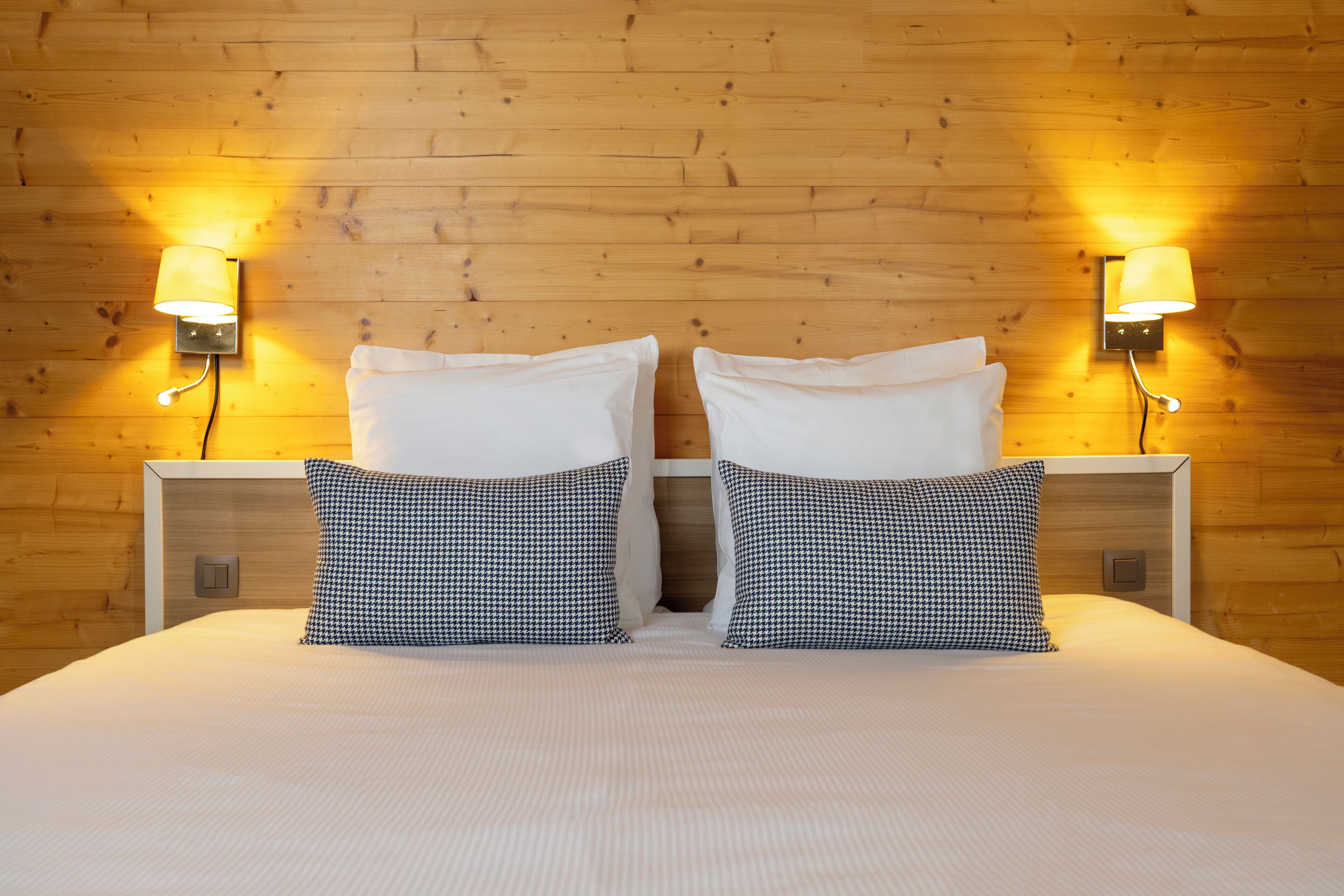 A neatly made bed with white linens and two houndstooth pillows, flanked by two wall-mounted lamps with a wooden backdrop.