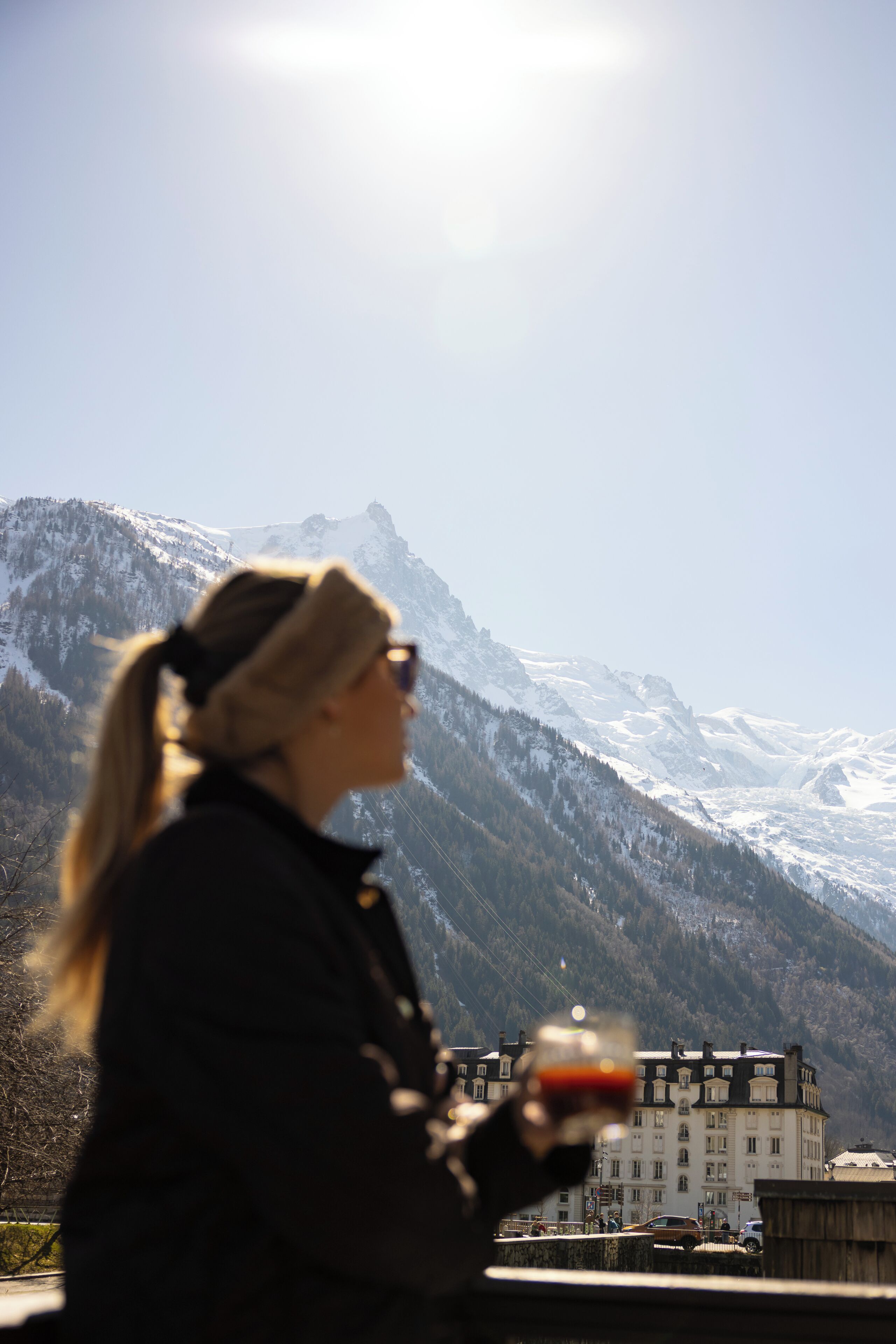 Una persona sosteniendo un vaso con una bebida, con vistas a un paisaje montañoso y un edificio al fondo.