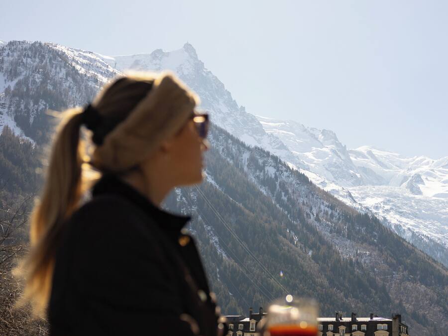 A person holding a glass with a beverage, overlooking a mountainous landscape with a building in the background.