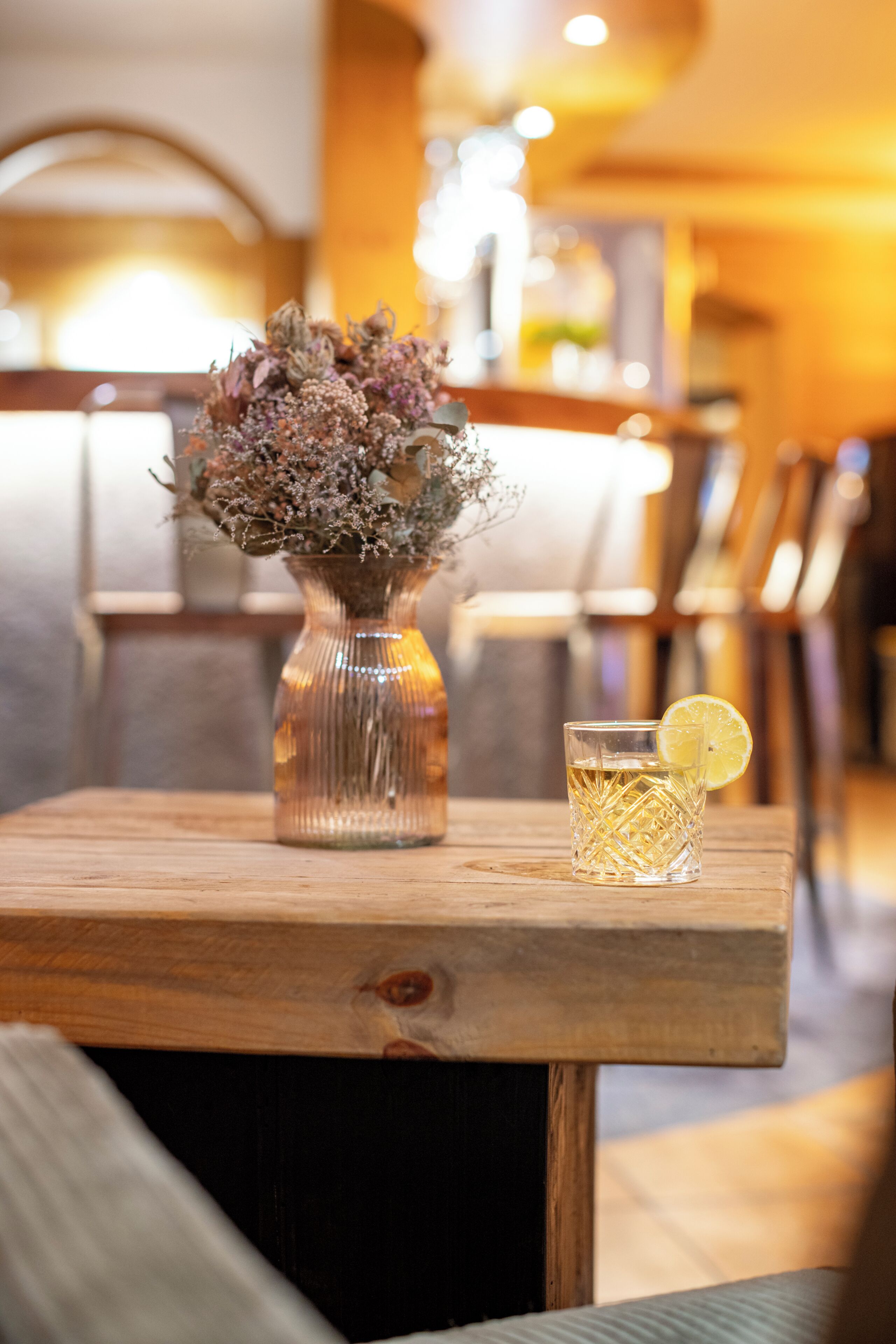 A wooden table with a vase of dried flowers and a glass of water with a lemon slice.