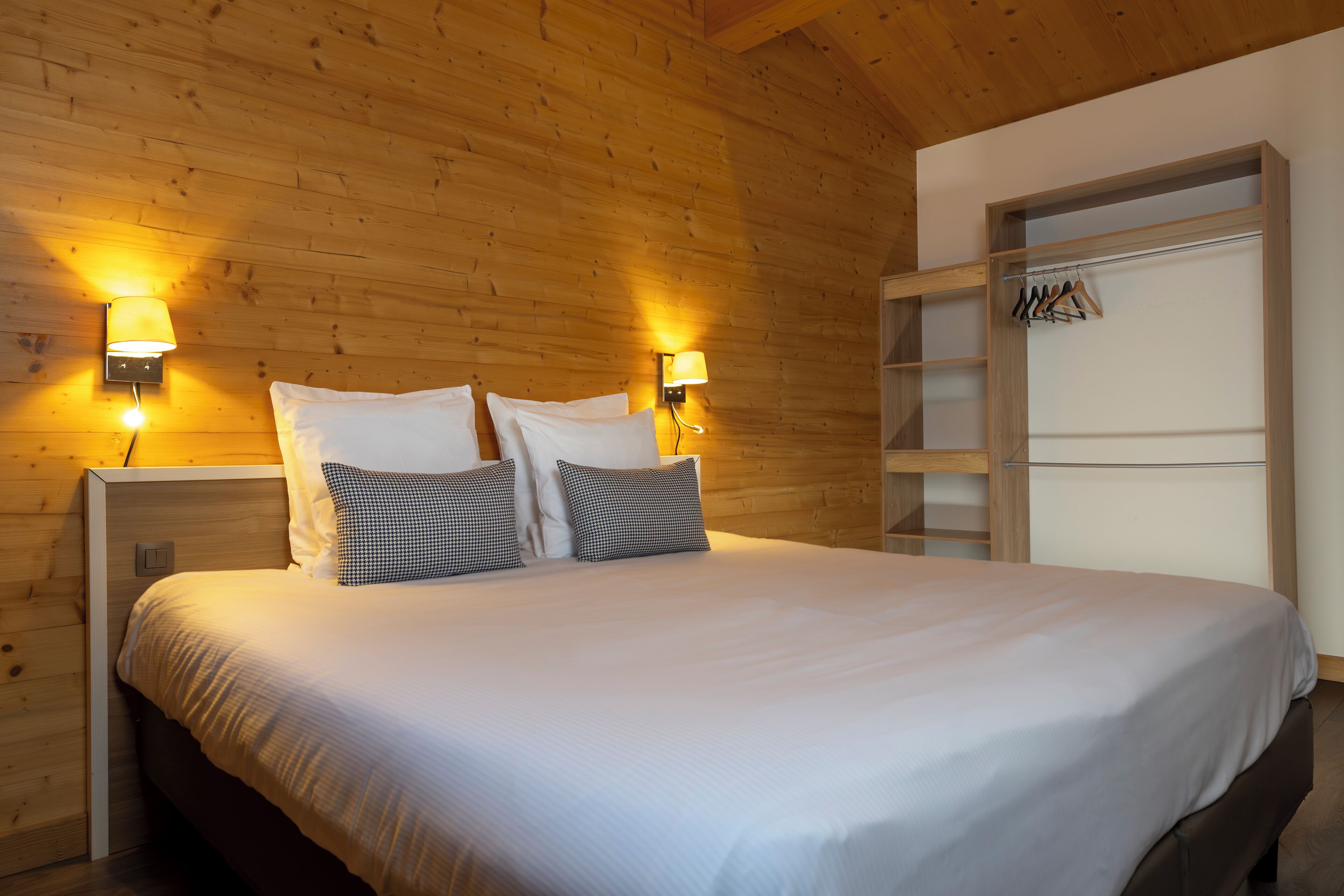 Una cama bien hecha con sábanas blancas y dos almohadas, paredes de madera y un armario de madera con espacio para colgar.