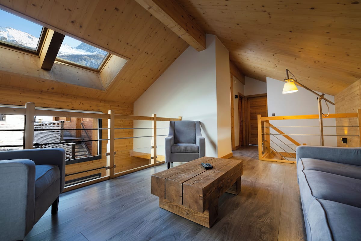 Een moderne loft-kamer met een schuine kap, houten balken, een groot raam met uitzicht op besneeuwde bergen, een grijze bank, een houten salontafel en een grijze fauteuil.