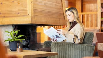 Une personne est assise dans une pièce chaleureuse aux murs en bois, avec une cheminée et une plante en pot, et elle est en train de lire un magazine.