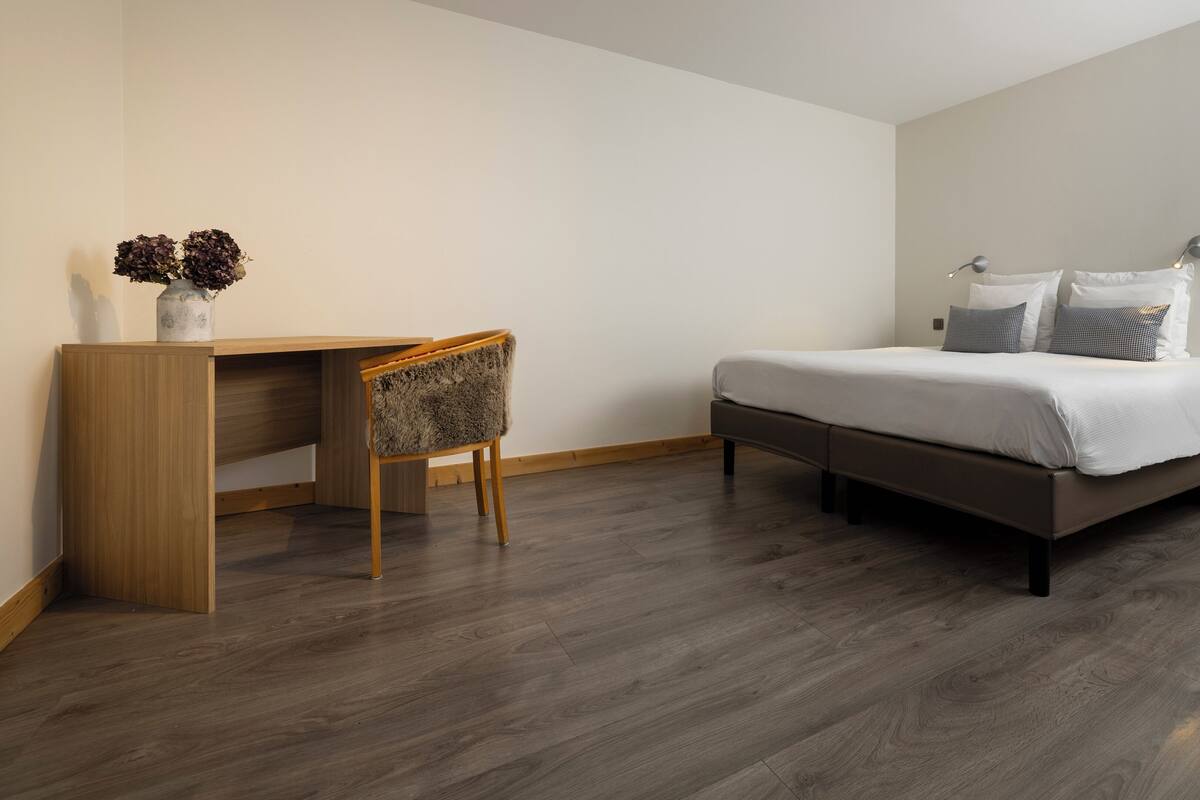 Een slaapkamer met een bed, nachtkastje, stoel en een bureau met een bloevas.