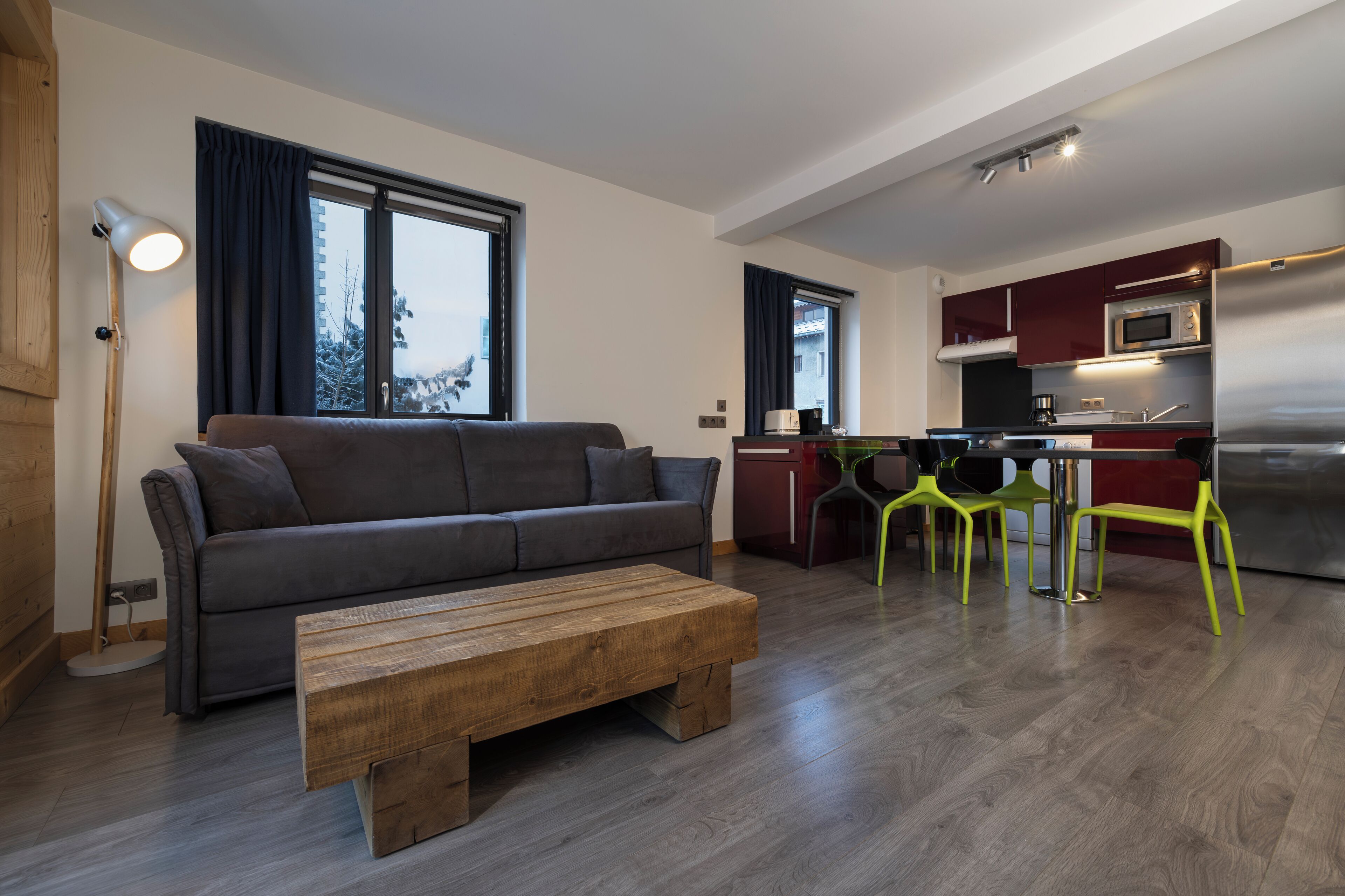 Una sala de estar moderna con un sofá gris, una mesa de centro de madera y sillas amarillo brillante. Se ve una cocina con electrodomésticos de acero inoxidable y armarios rojos al fondo.