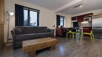 Una sala de estar moderna con un sofá gris, una mesa de centro de madera y sillas amarillo brillante. Se ve una cocina con electrodomésticos de acero inoxidable y armarios rojos al fondo.