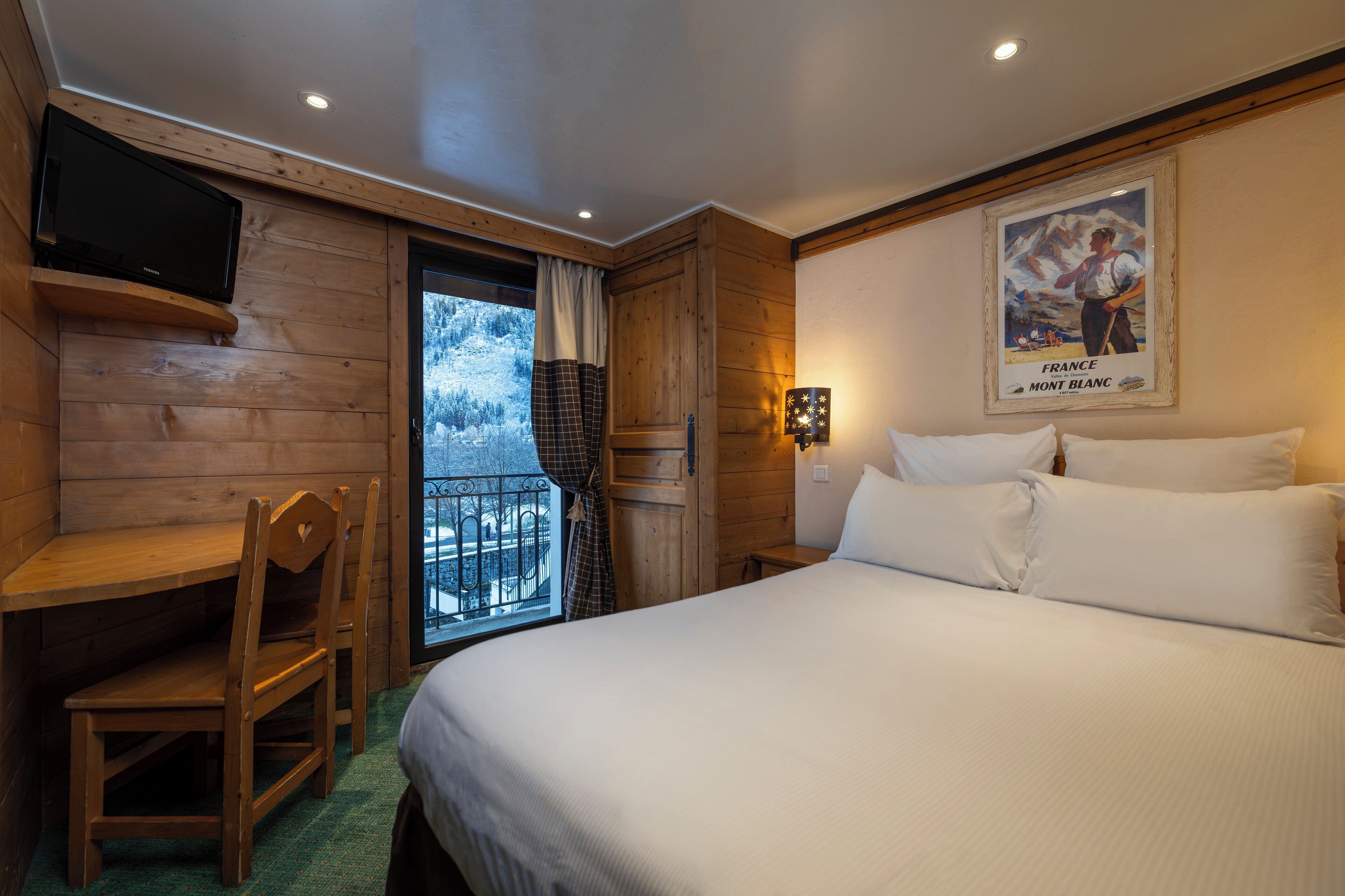 Quarto de hotel com decoração em madeira, cama com roupa branca, escrivaninha com cadeira, televisão na parede e um quadro na parede.
