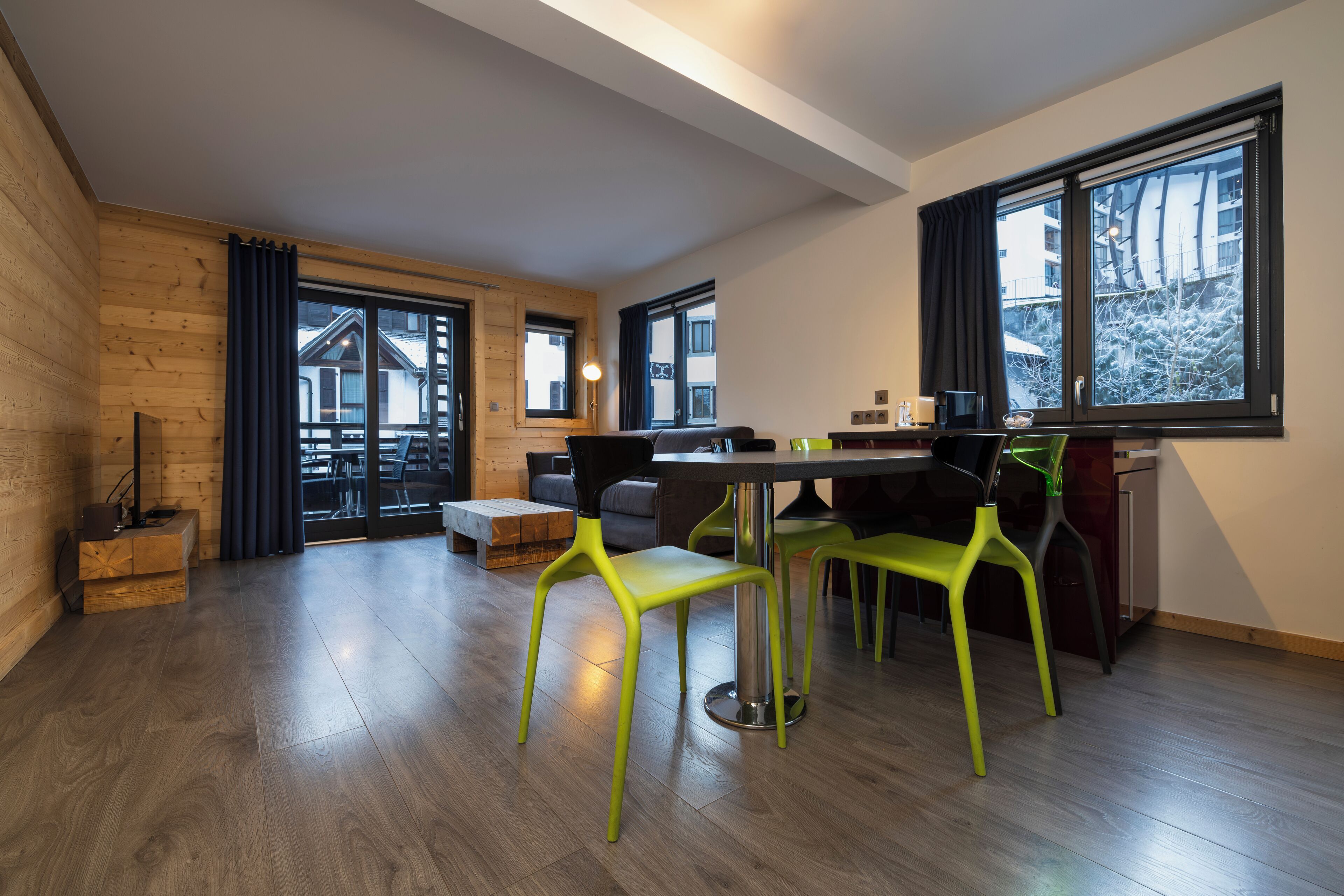 Un ambiente moderno con pavimento in legno, una zona pranzo con tavolo e sedie e ampie finestre con tende.