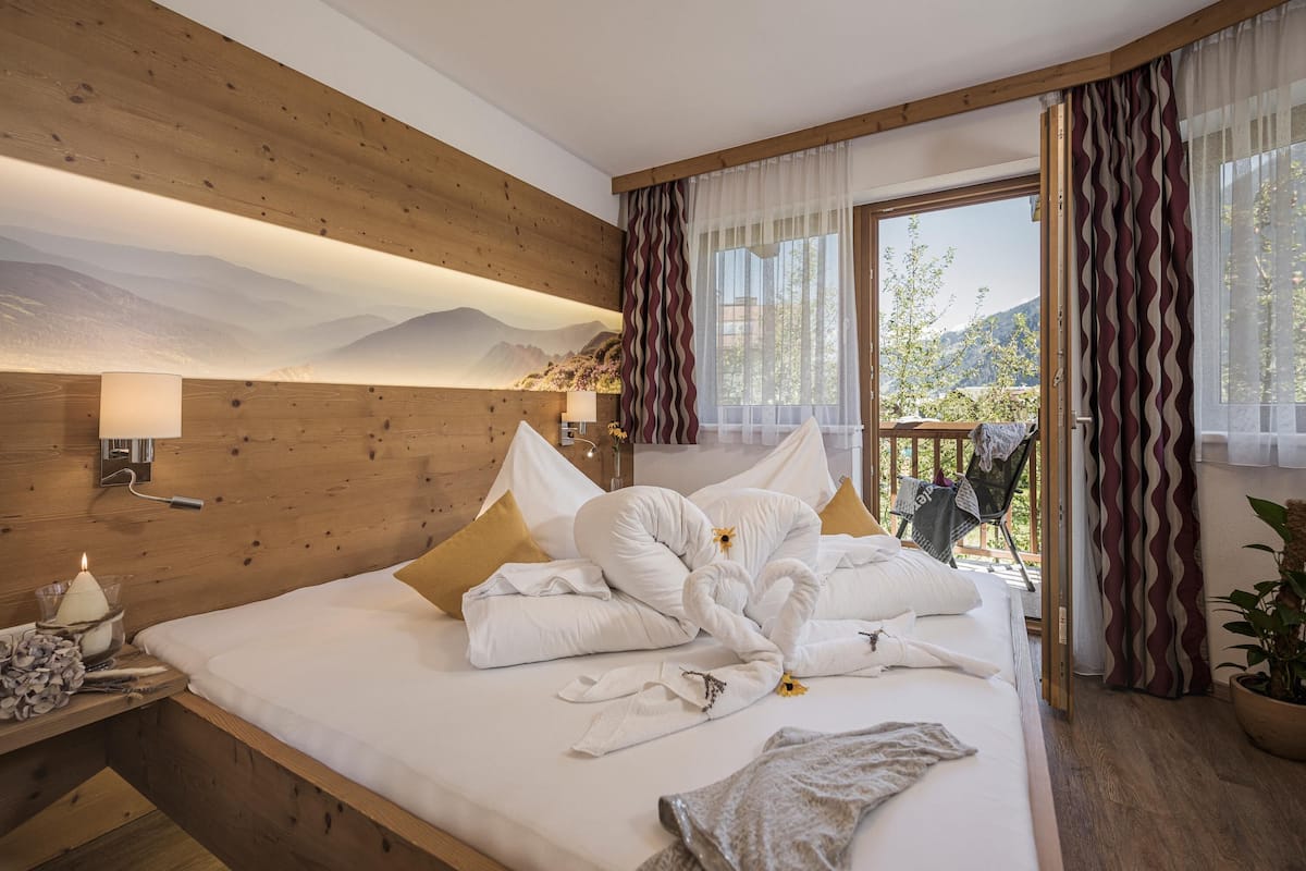 Une chambre à coucher avec une tête de lit en bois, un lit recouvert de draps blancs et une vue sur les montagnes par une fenêtre ornée de rideaux.