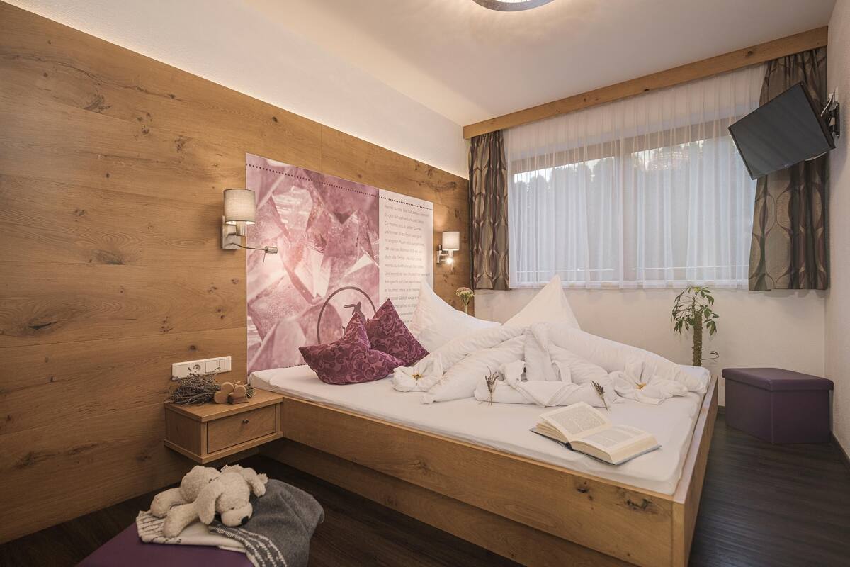 Une chambre à coucher avec une tête de lit en bois, un lit recouvert de draps blancs, une table de chevet avec une lampe, une fenêtre ornée de rideaux légers et un téléviseur fixé au mur.