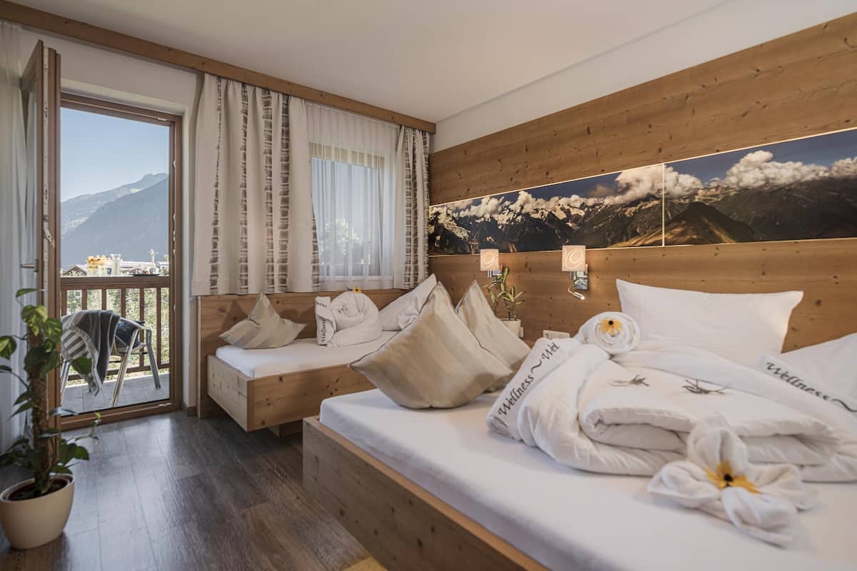 Une chambre d’hôtel avec une tête de lit en bois, une grande fenêtre avec des rideaux, un lit avec du linge de lit blanc et des serviettes, une petite table de chevet et une vue sur les montagnes.