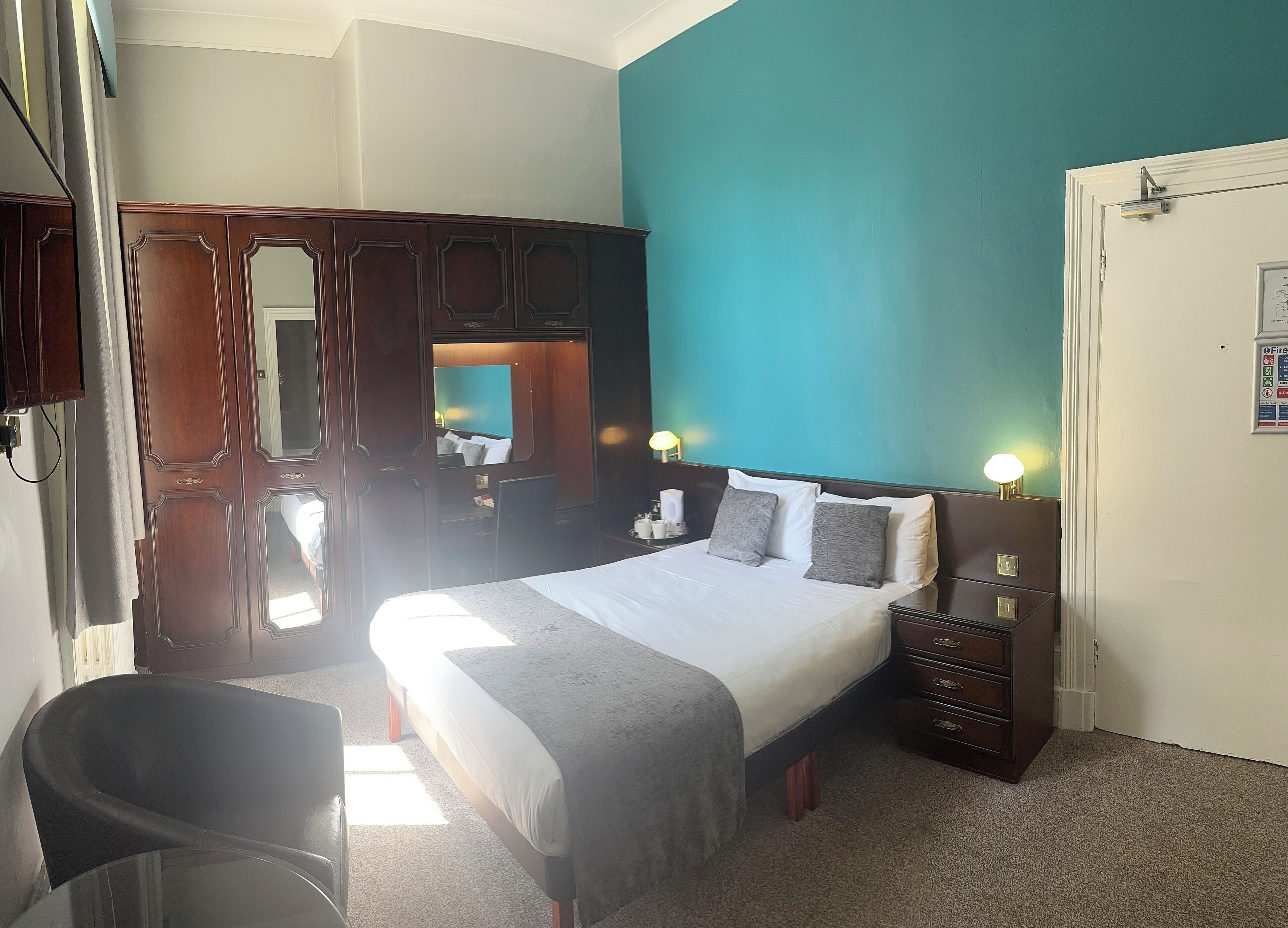 A hotel room with a large bed, a wooden wardrobe, a desk with a lamp, and a chair.
