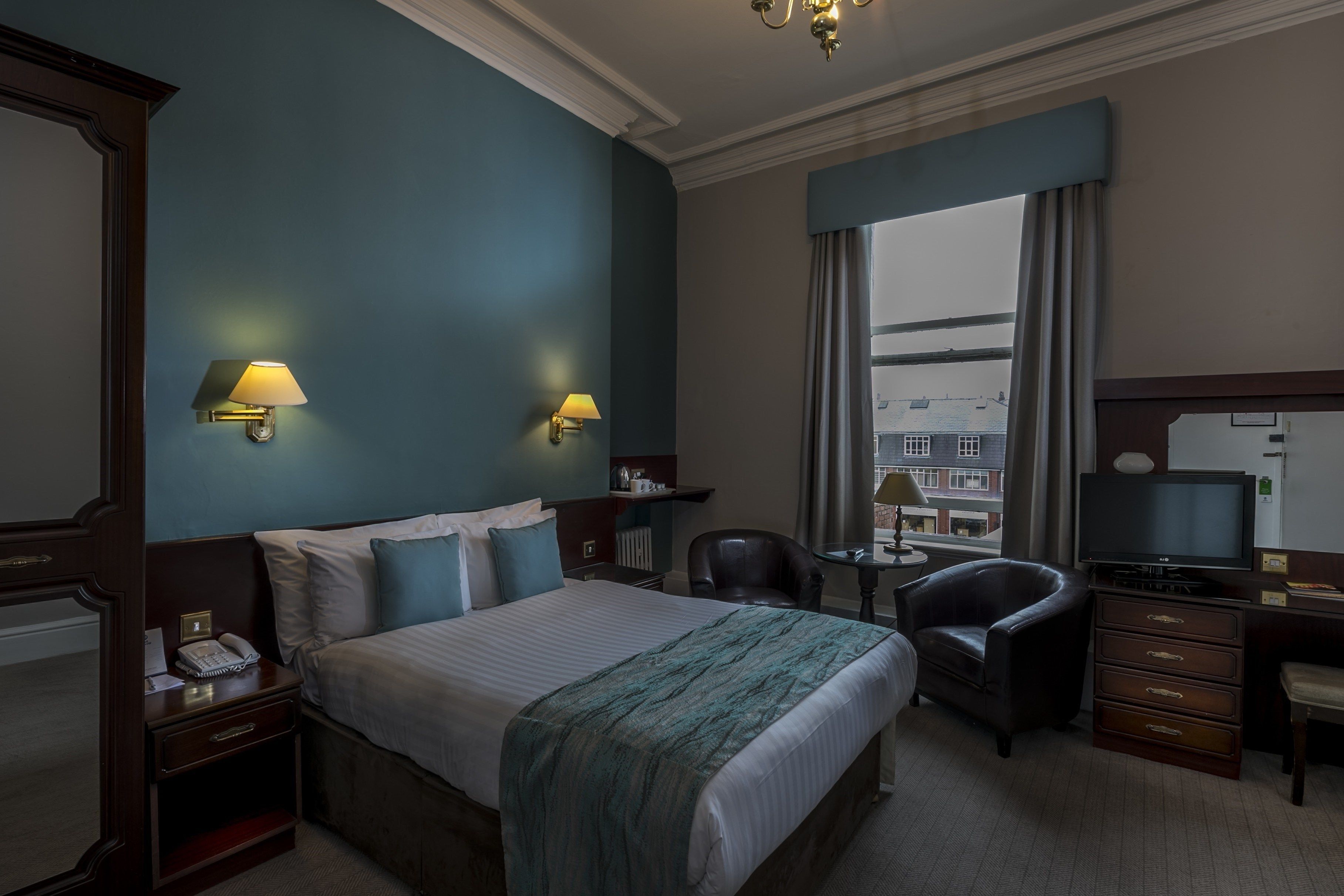 A hotel room with a large bed, two bedside lamps, a desk with a chair, a television, and a view of the city through the window.