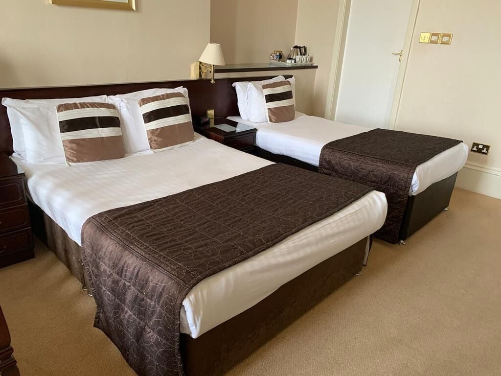 A hotel room with two beds, each with a brown bedspread and white linens. There is a nightstand with a lamp and a small table with a coffee maker.