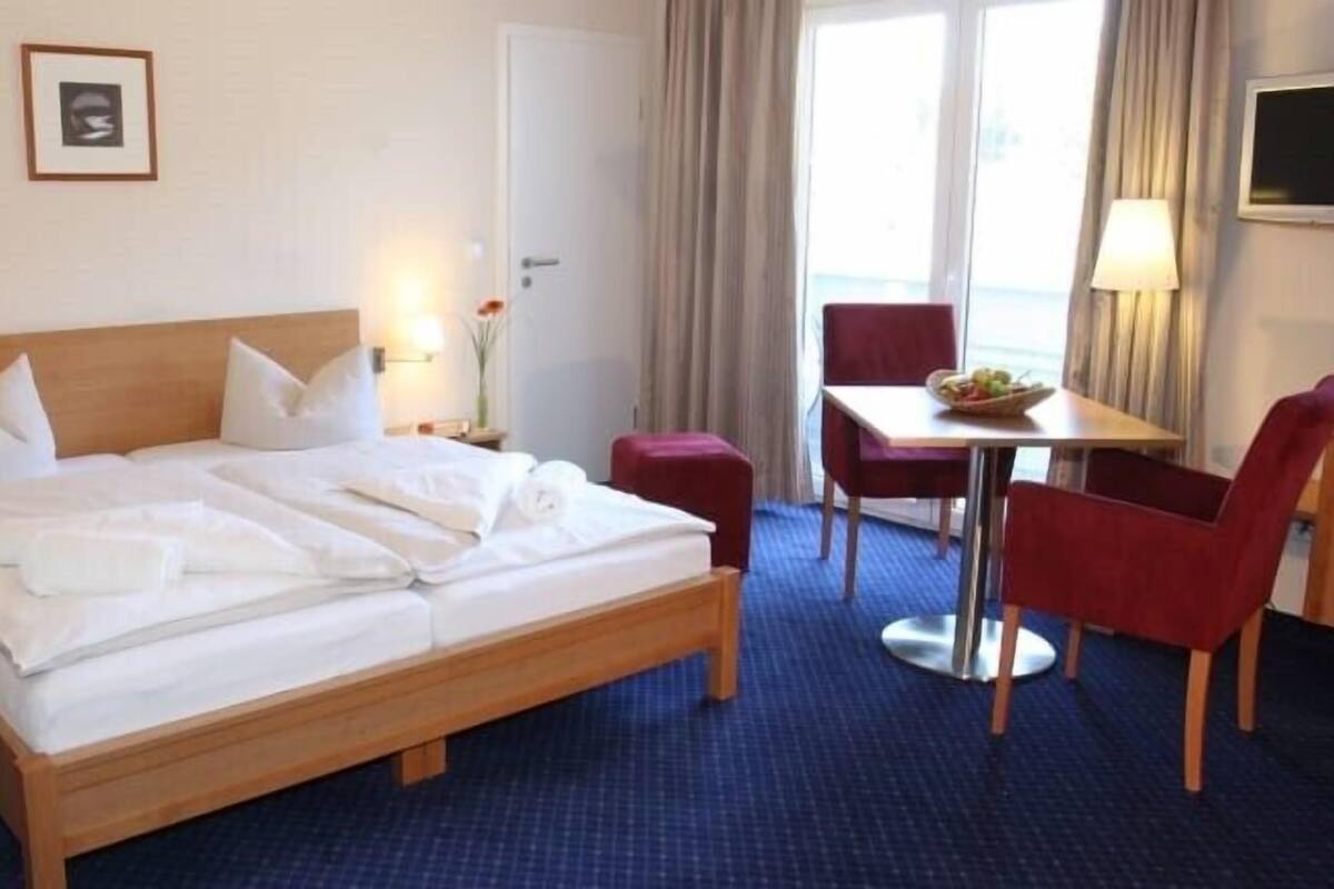 Une chambre d’hôtel avec un grand lit, un bureau avec deux chaises, une fenêtre avec des rideaux et un tableau encadré au mur.