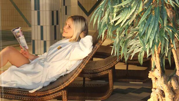 Una persona se relaja en una silla de mimbre dentro de un espacio luminoso con suelo de baldosas y una planta en maceta cerca.