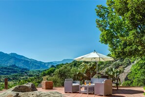 Une terrasse extérieure aménagée avec des meubles en osier, une table et un grand parasol, offrant une vue sur un paysage montagneux.