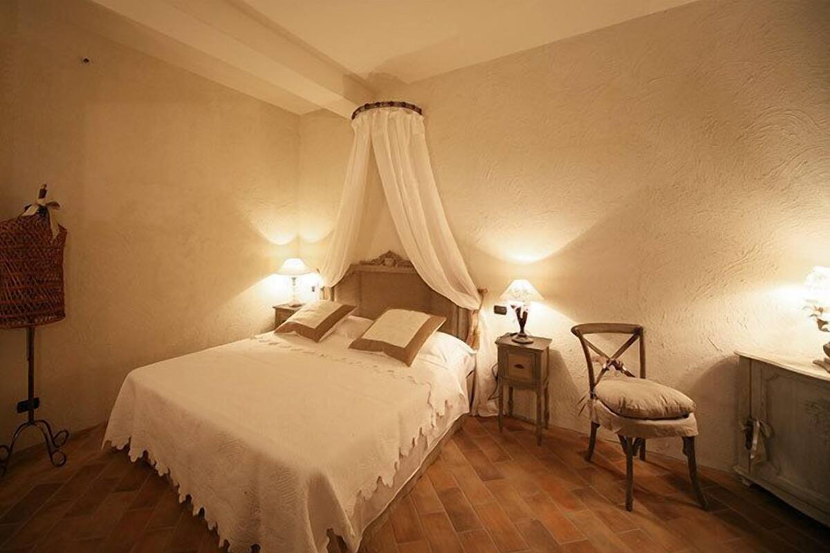 Een slaapkamer met hemelbed, nachtlampjes, een stoel en een klein tafeltje.